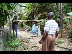 A resident, reportedly the grandfather of one of the victims, looks on as a policeman monitors the cordoned-off crime scene in Hashburn, off Brooks Level Road in Stony Hill. 