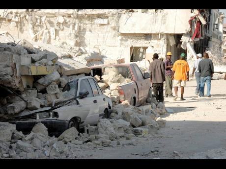Survivors of the 2010 Haiti earthquake walk by motor vehicles crushed by a collapsed building shortly after the disaster which claimed hundreds of thousands of lives in that country 10 years ago.