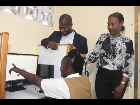 Father Carl F. Clarke (left), principal at the Holy Trinity High School, and Gail Moore (right), partner and environment lead at PwC Jamaica, catch up with grade nine student Krianna Gilzene, during set up of  one of eight newly donated desktop computers for the enrichment centre that hosts the institution’s academic intervention programme. 