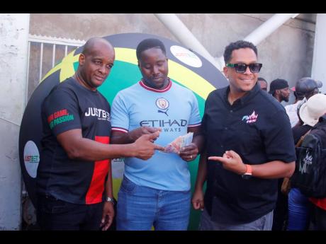 Credit: Contributed JustBet Product Manager Chris Sawyers (left) and FYAH 105 host Bambino (right) share in the winning moment with Bebito Brown during the Ballin with JustBet outside broadcast at the Half-Way Tree Transport Centre in St Andrew.